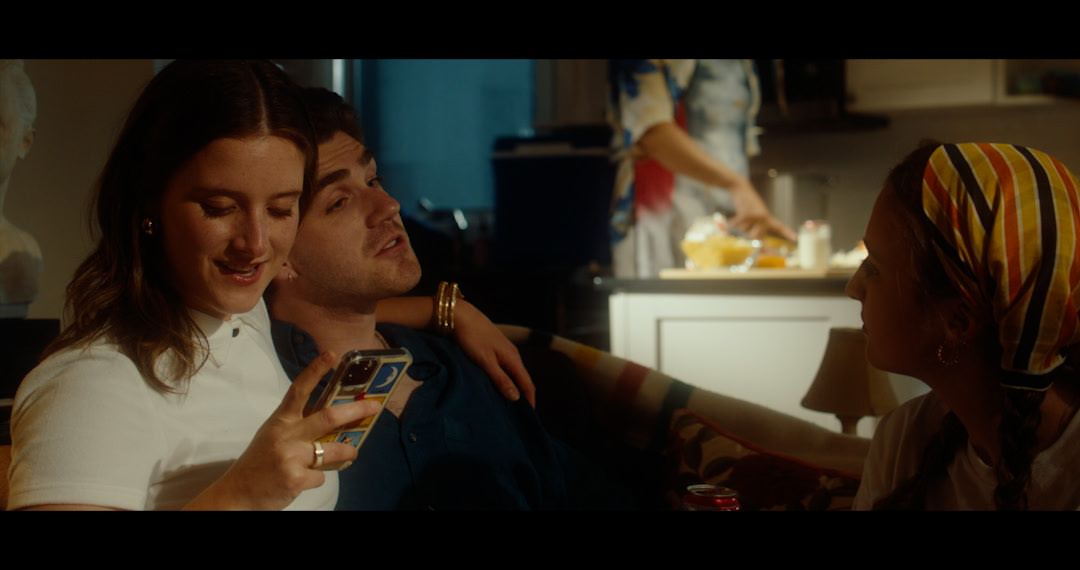 a couple is sitting around the couch with their hands around eachother. The woman is gazing at her phone while the man is chatting with a person. Behind them is a kitchen where someone appears to be chopping up ingredients.